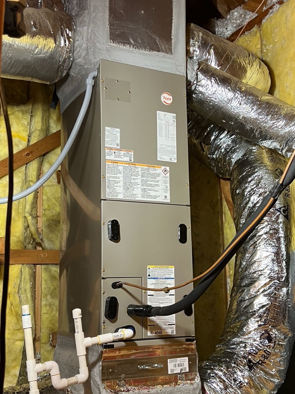 An HVAC air handler unit installed in an attic, connected to silver insulated flexible ductwork and white PVC drain pipes.