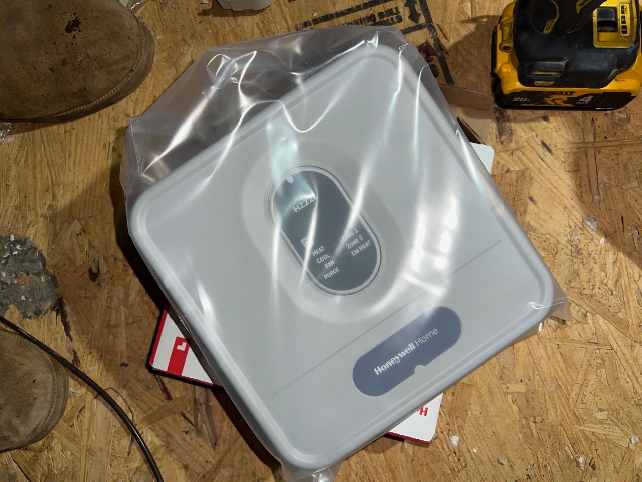 A light gray square device inside a clear plastic bag resting on a wooden floor near tools.
