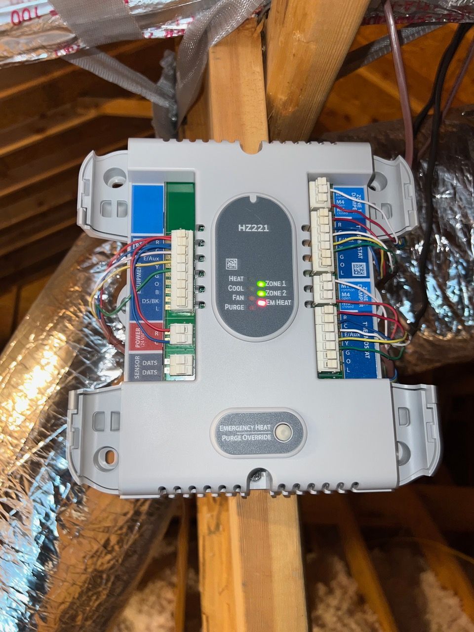 A Honeywell zoning control board with wired connections mounted on a wooden attic support beam, showing status LEDs.