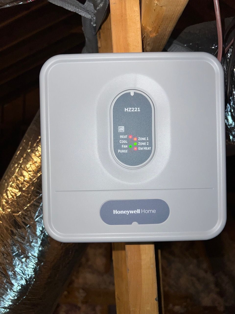A Honeywell EVC monitoring device mounted on a wooden beam in an attic with status lights illuminated.