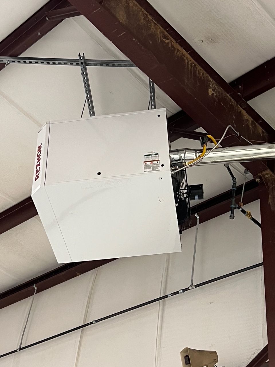 A white industrial ceiling-mounted unit heater suspended from metal roof beams in a large workshop or garage.