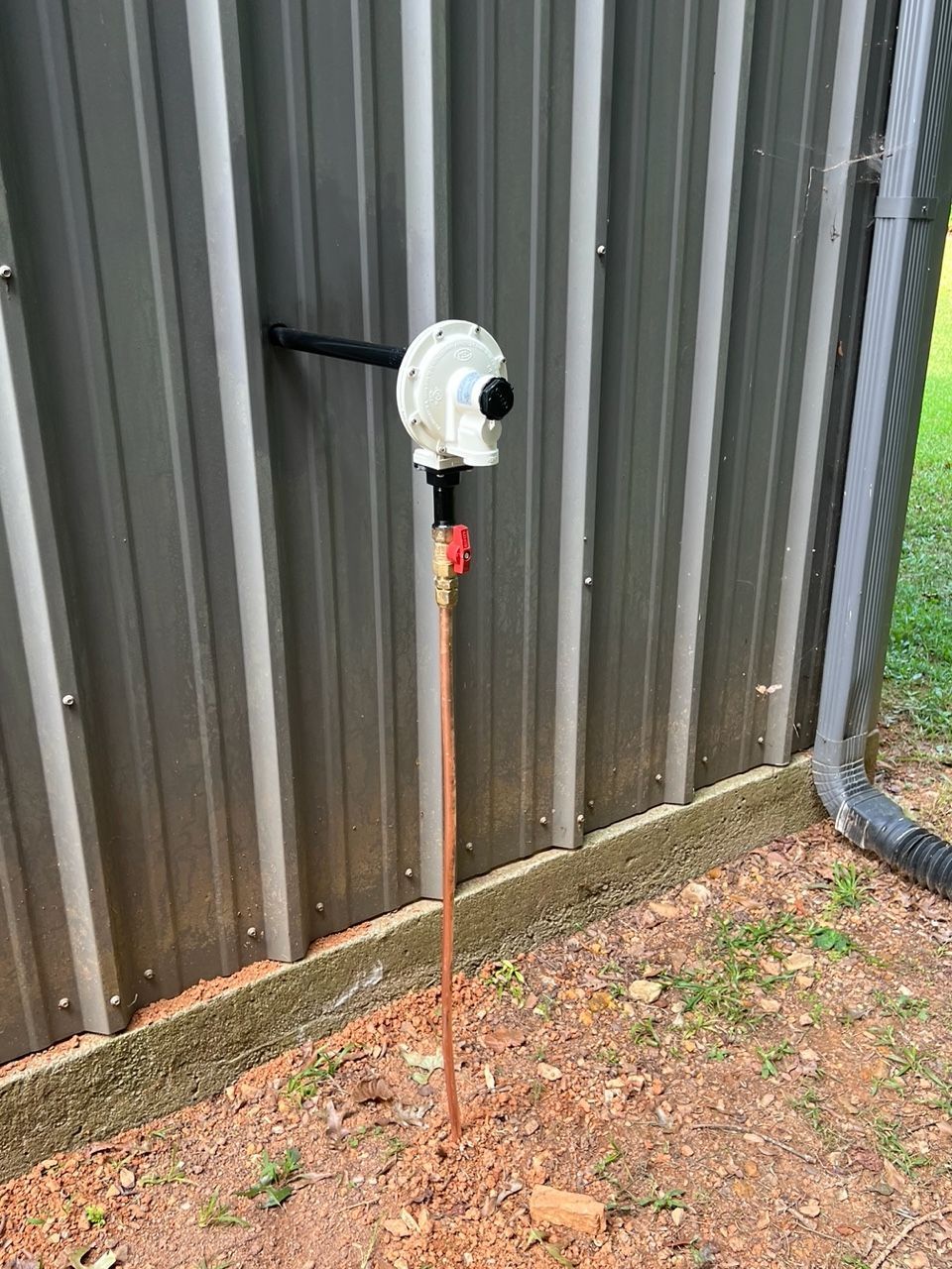 A white propane regulator attached to a metal building wall with a red-handled shutoff valve and copper supply line.