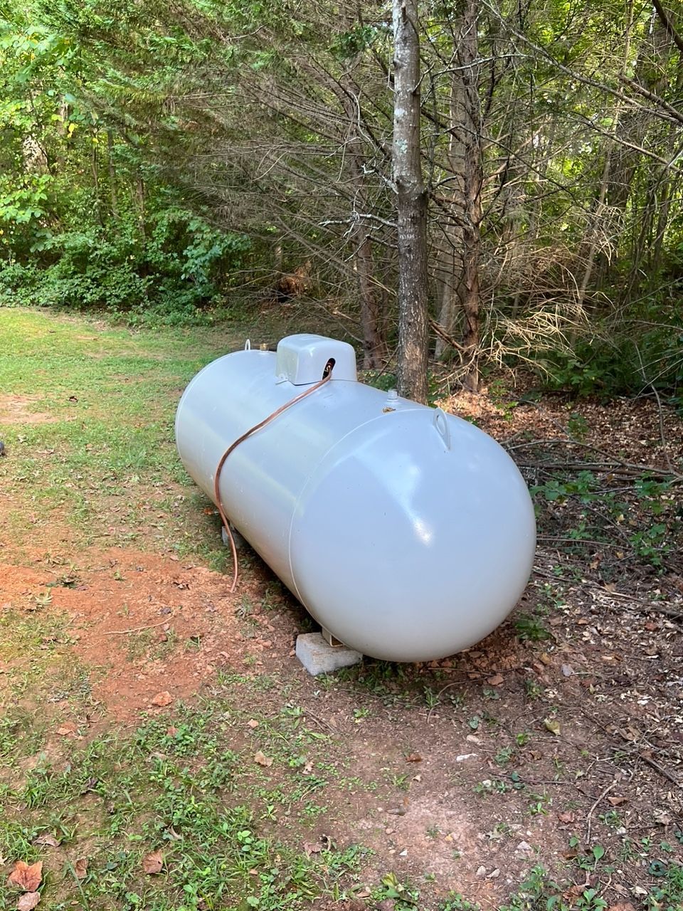 A light-gray propane tank rests on concrete blocks at the edge of a wooded area.