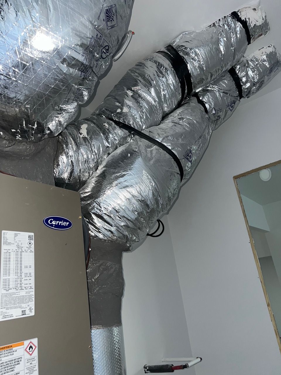 Silver, insulated flexible air ducts connected to the top of a Carrier HVAC unit in a room.
