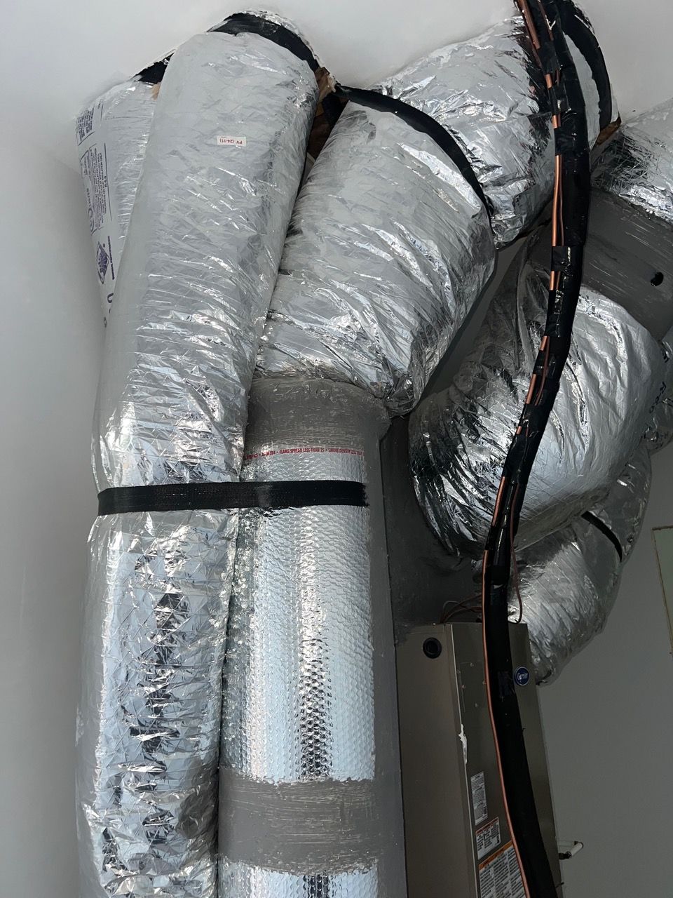 Silver-insulated flexible HVAC ductwork attached to a ceiling-mounted unit with black straps and wiring.