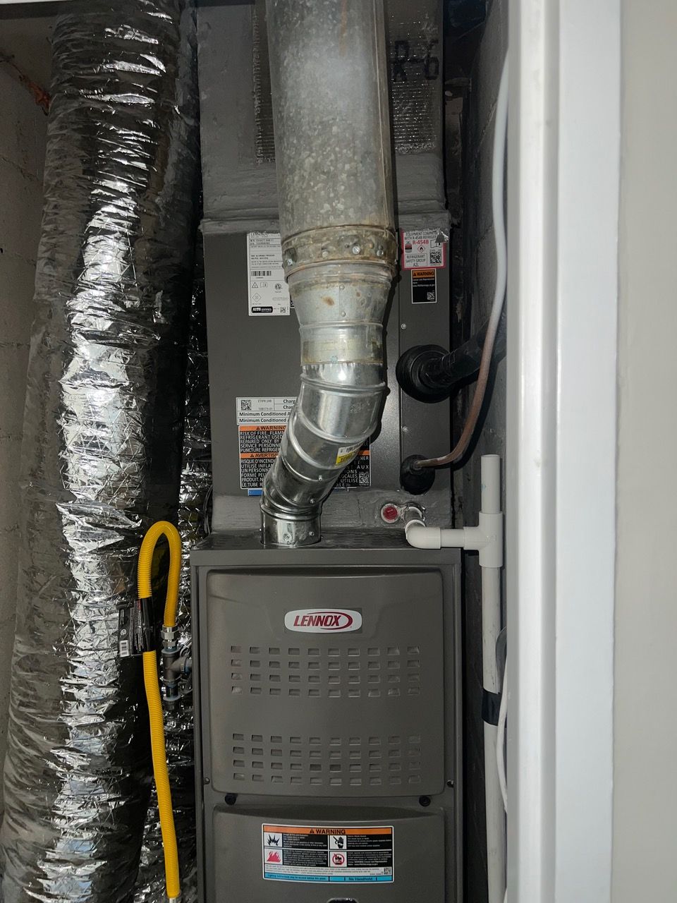 A grey Lennox furnace unit with a metal exhaust pipe and silver insulated ductwork installed in a utility closet.