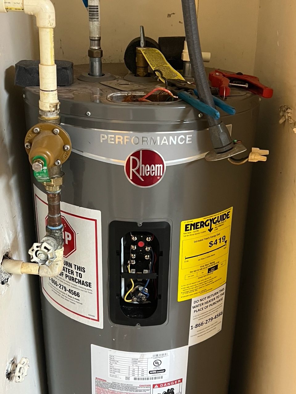 A gray Rheem Performance electric water heater installed in a corner with a pressure valve and tools on top.