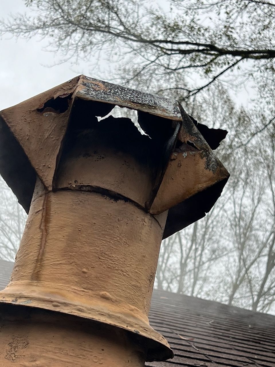 A damaged, weathered metal chimney cap on a roof with a cloudy, tree-lined background.