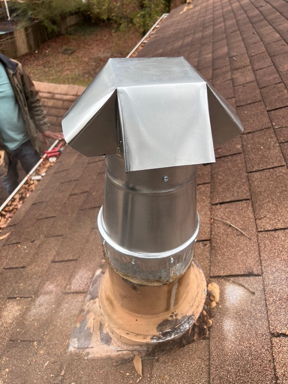 A metal chimney cap installed on a roof, with a portion of a person visible in the background near the gutter.