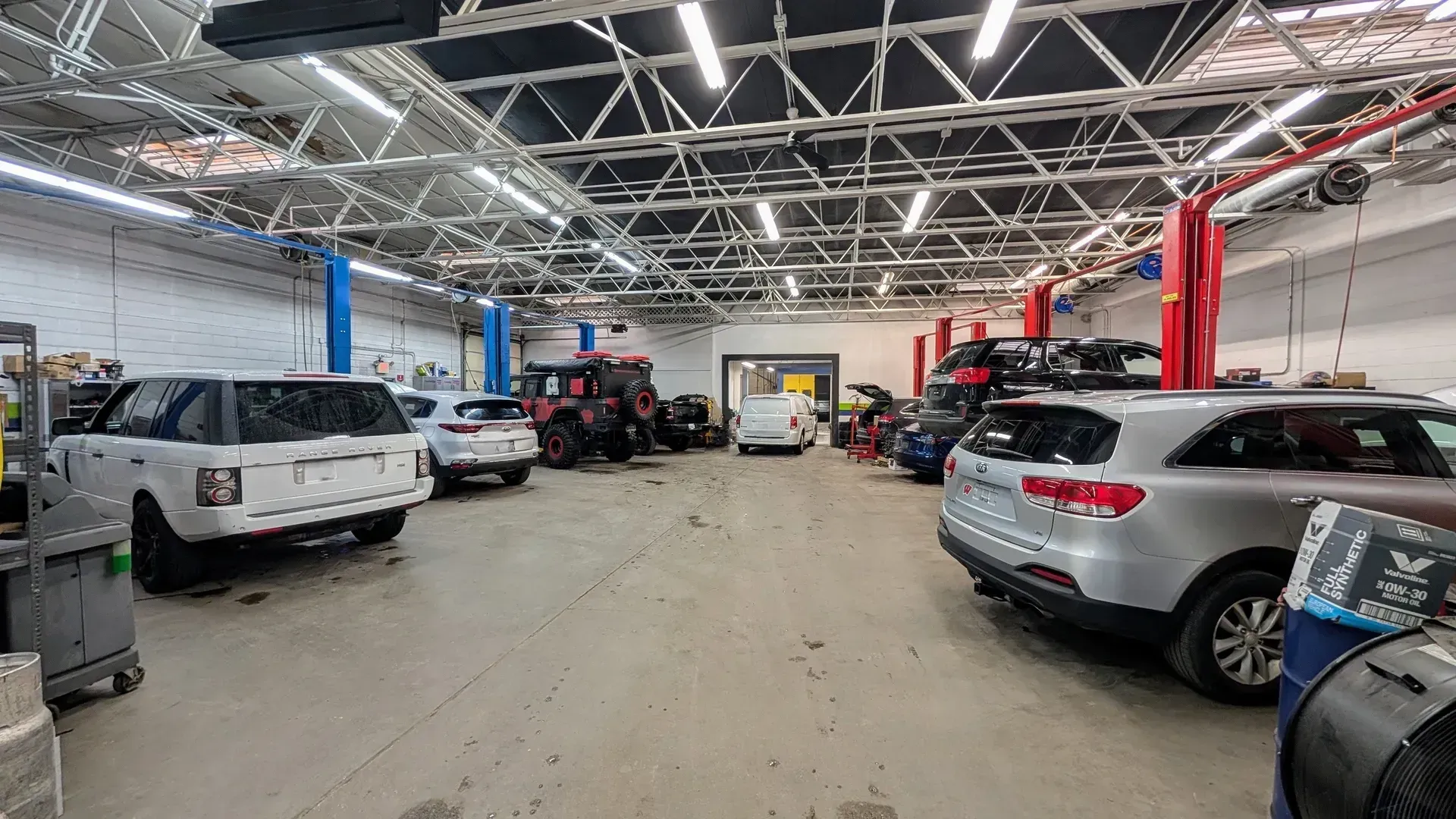 A bright auto repair shop interior featuring several cars, including white and silver SUVs, under a metal roof structure.