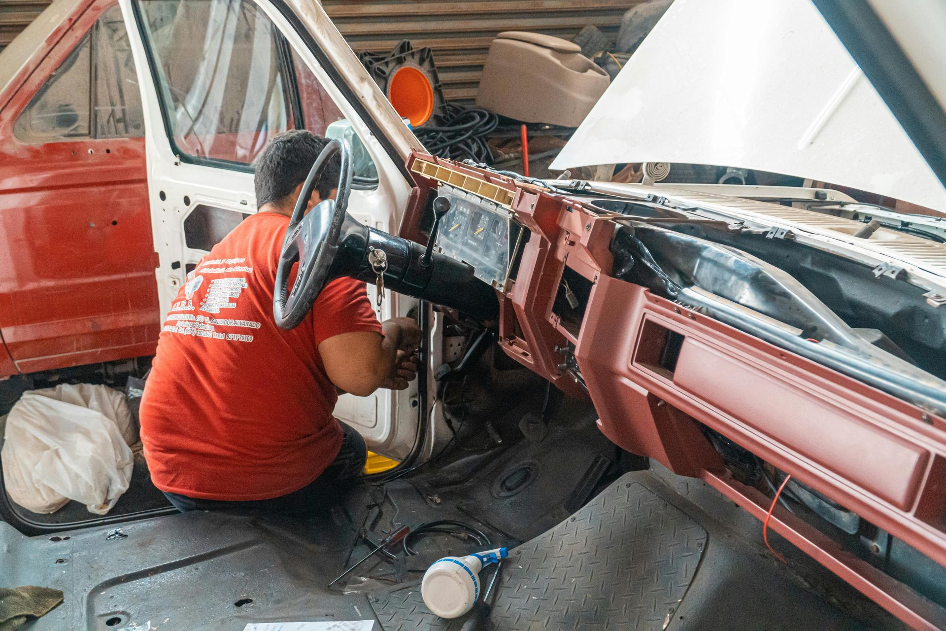 A person in an orange shirt works on the interior wiring and dashboard of a vehicle under repair in a garage.