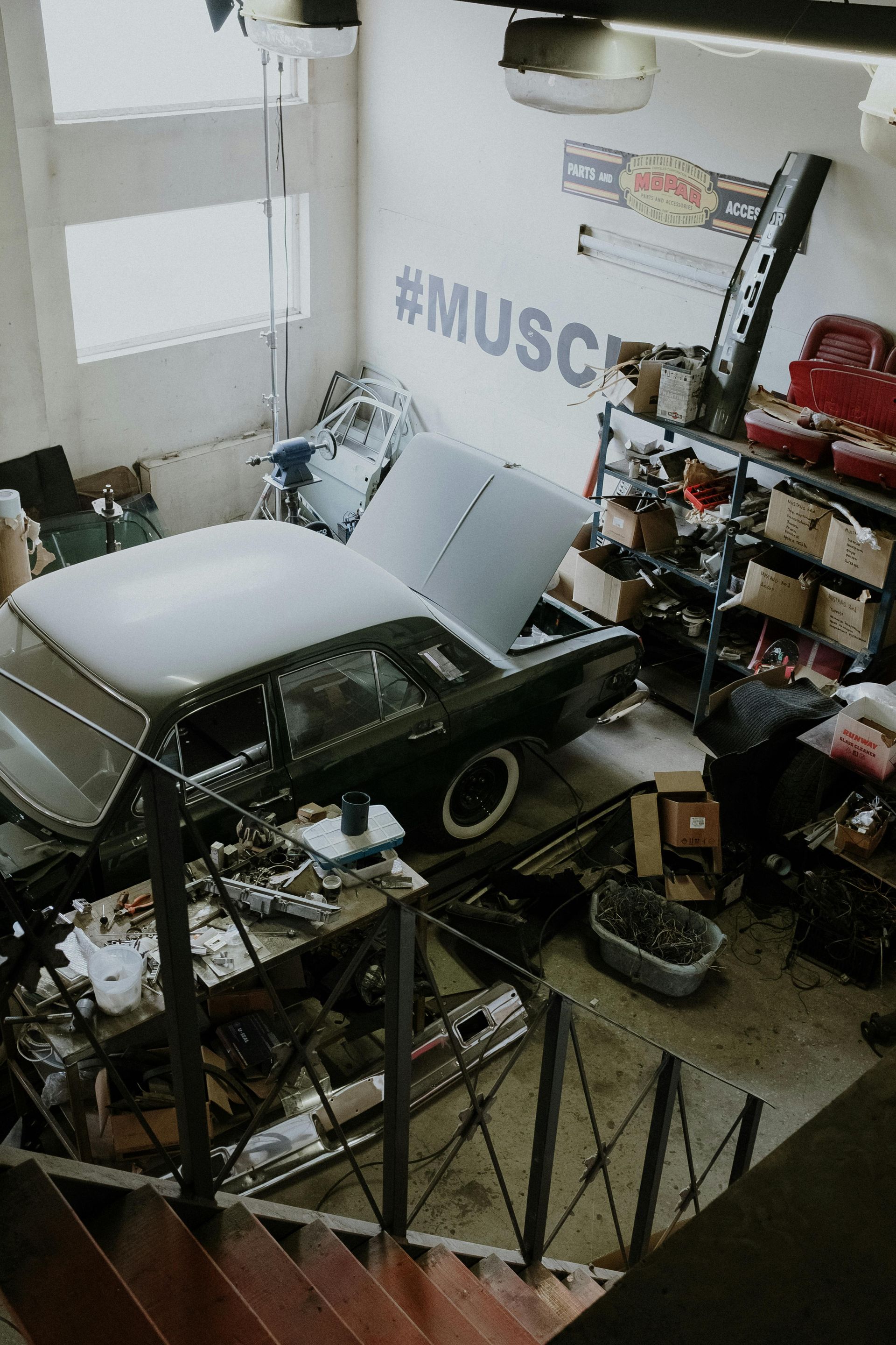 A high-angle view of a workshop with a dark green car under repair, metal shelving, tools, and a