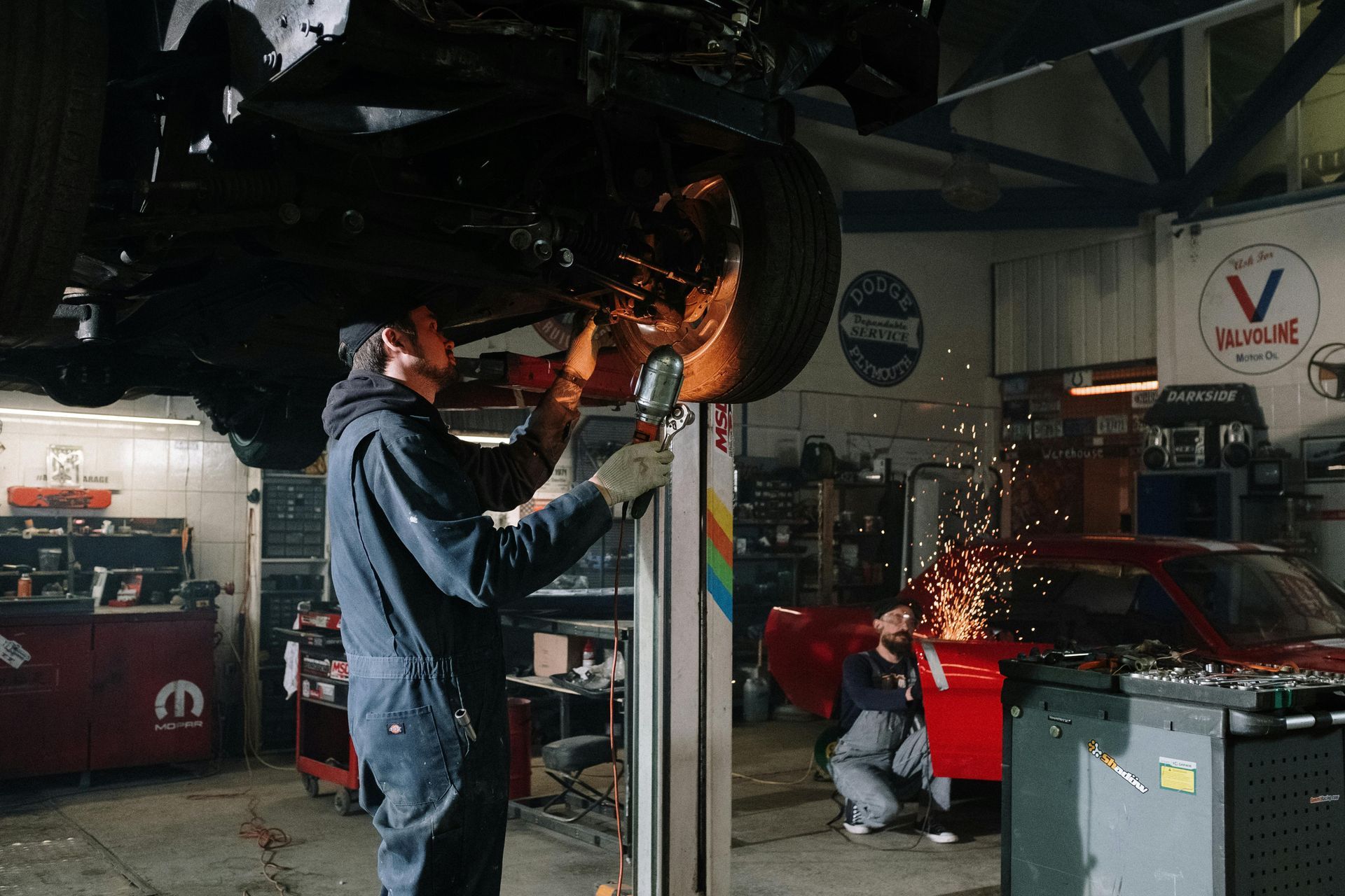 A mechanic uses a power tool on a lifted car in a garage while another person works on a red vehicle nearby.
