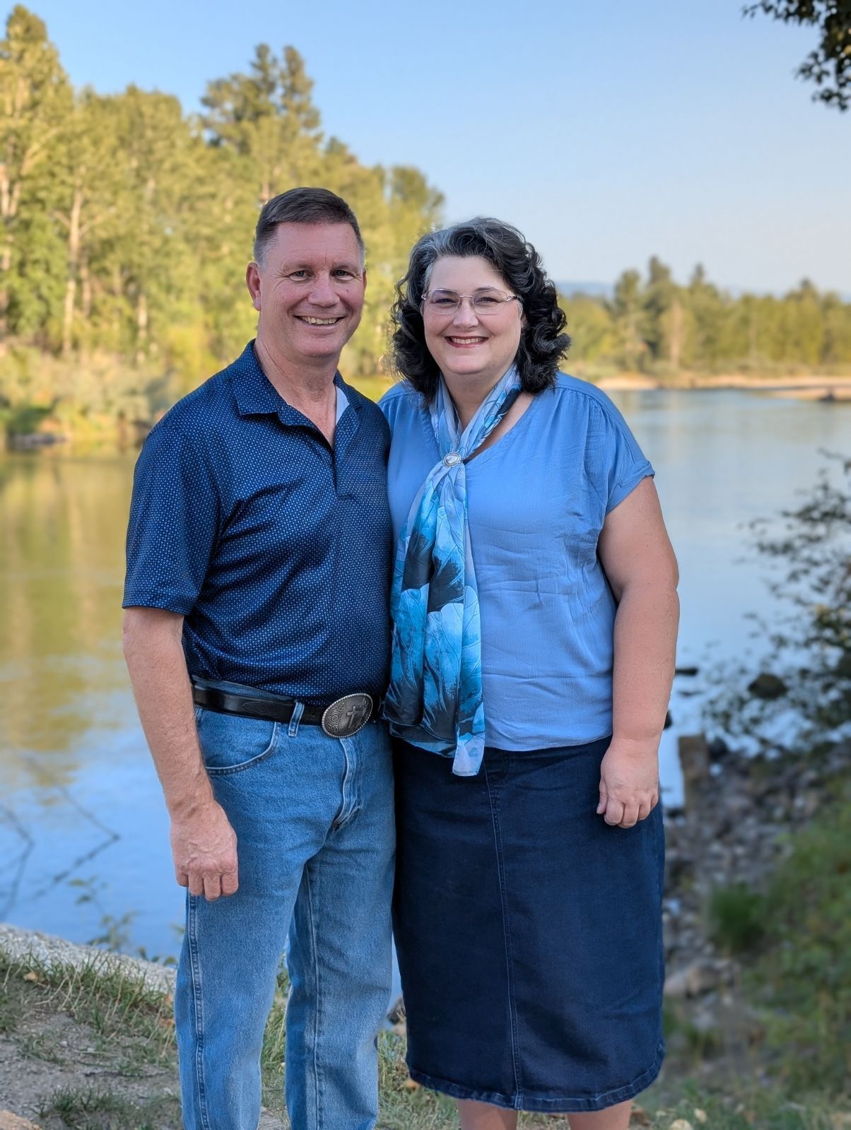 a man and a woman are posing for a picture in the woods . the man is wearing a cowboy hat .