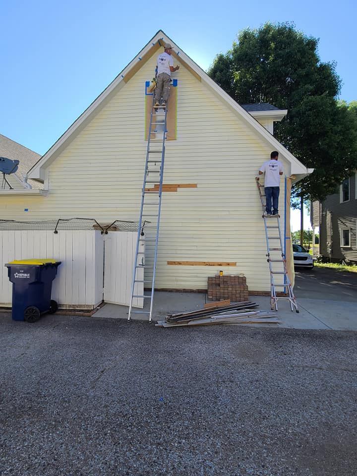 Two workers on ladders painting a beige house exterior beside a tree and trash bin