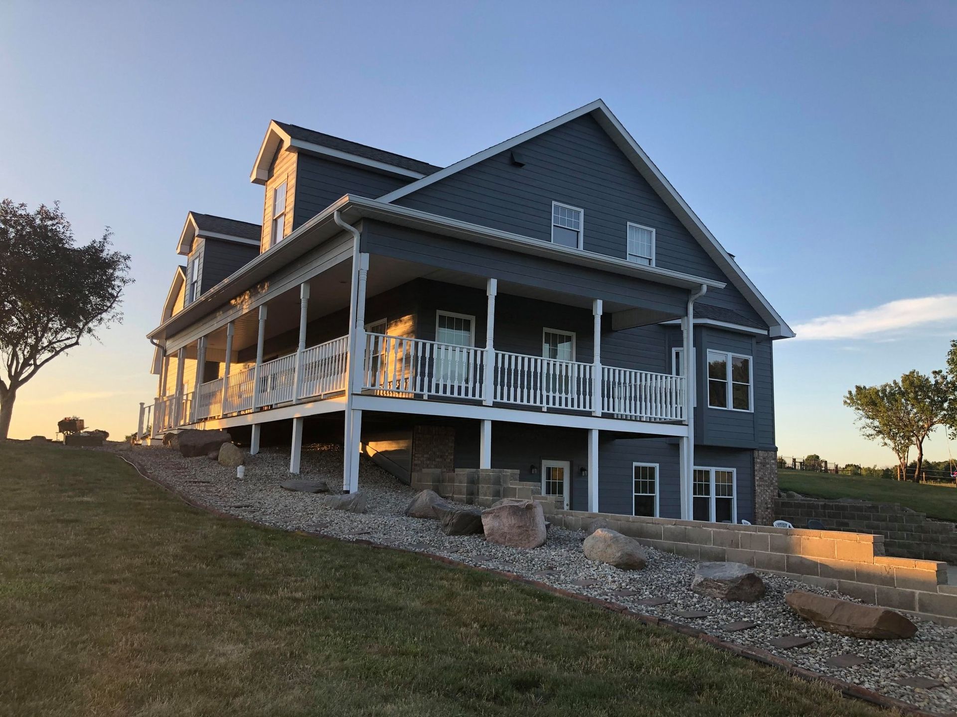 Large gray farmhouse with a wraparound porch on a grassy hillside at sunset