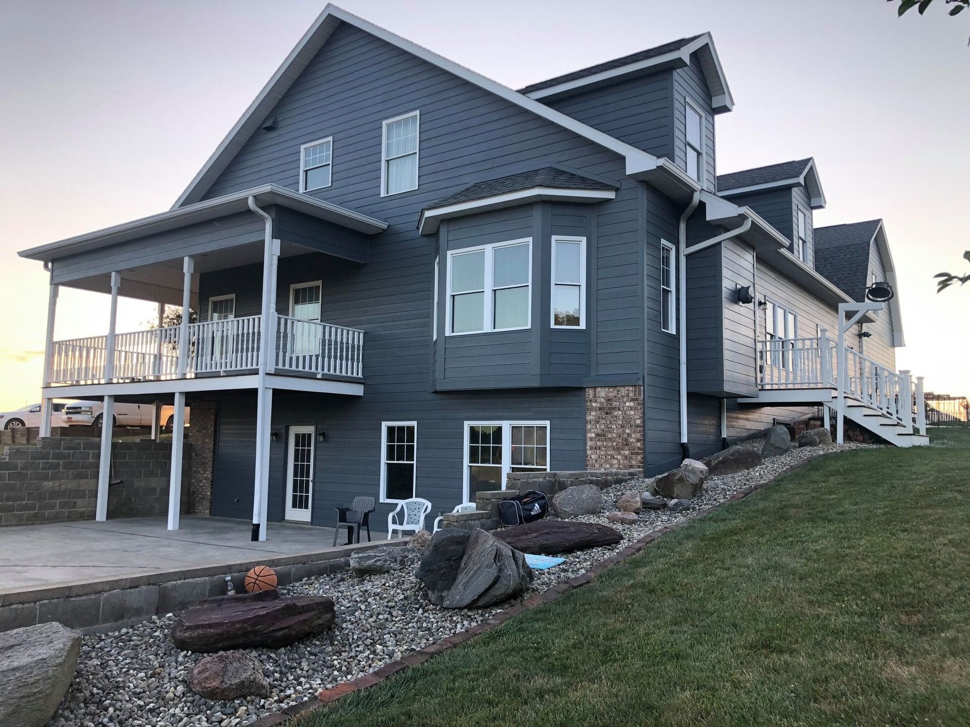 Gray two-story house with white trim, wraparound porch, and stone foundation on a grassy slope.
