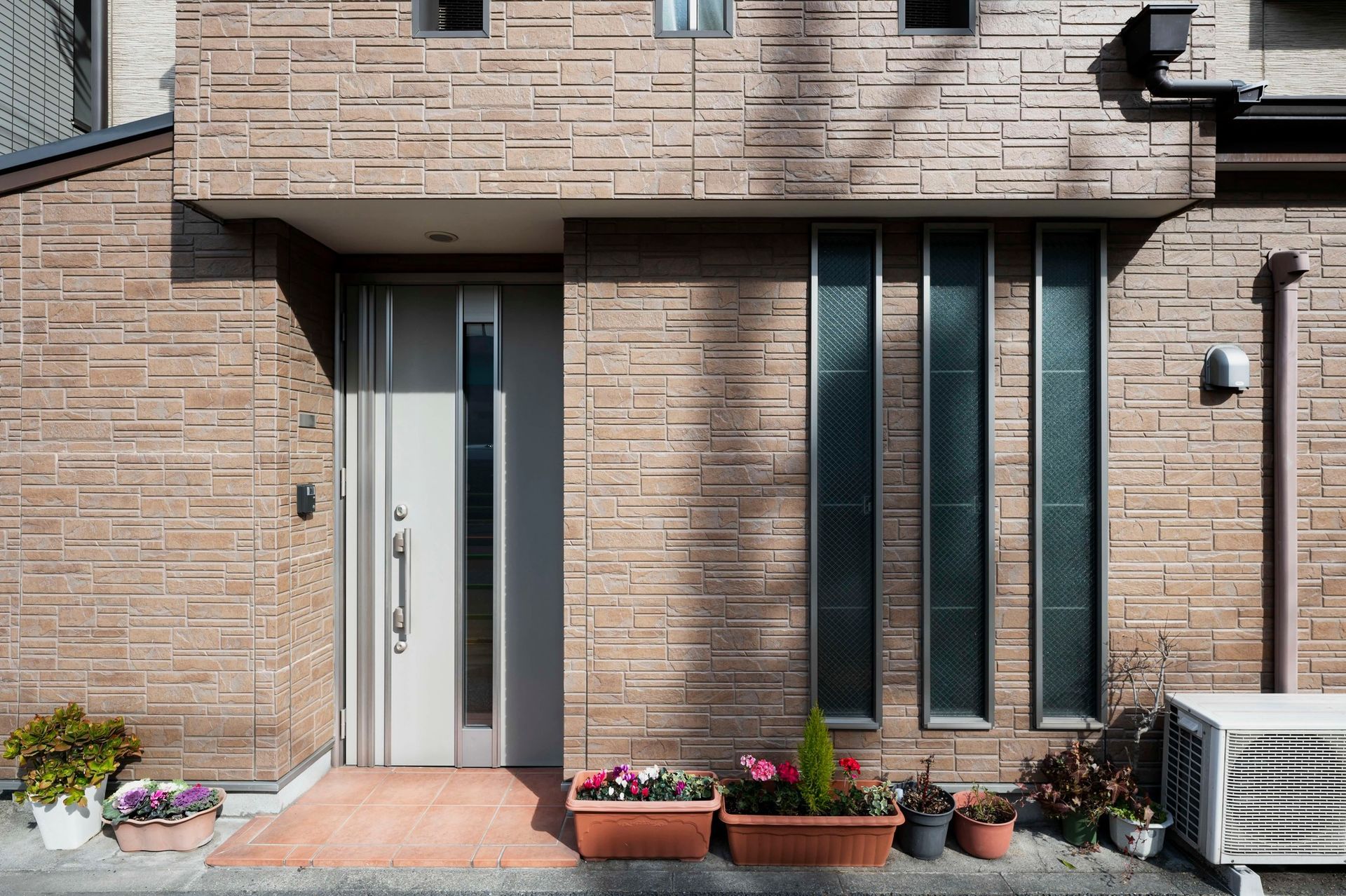 Modern brick townhouse entrance with white door, tall windows, potted flowers, and outdoor AC unit