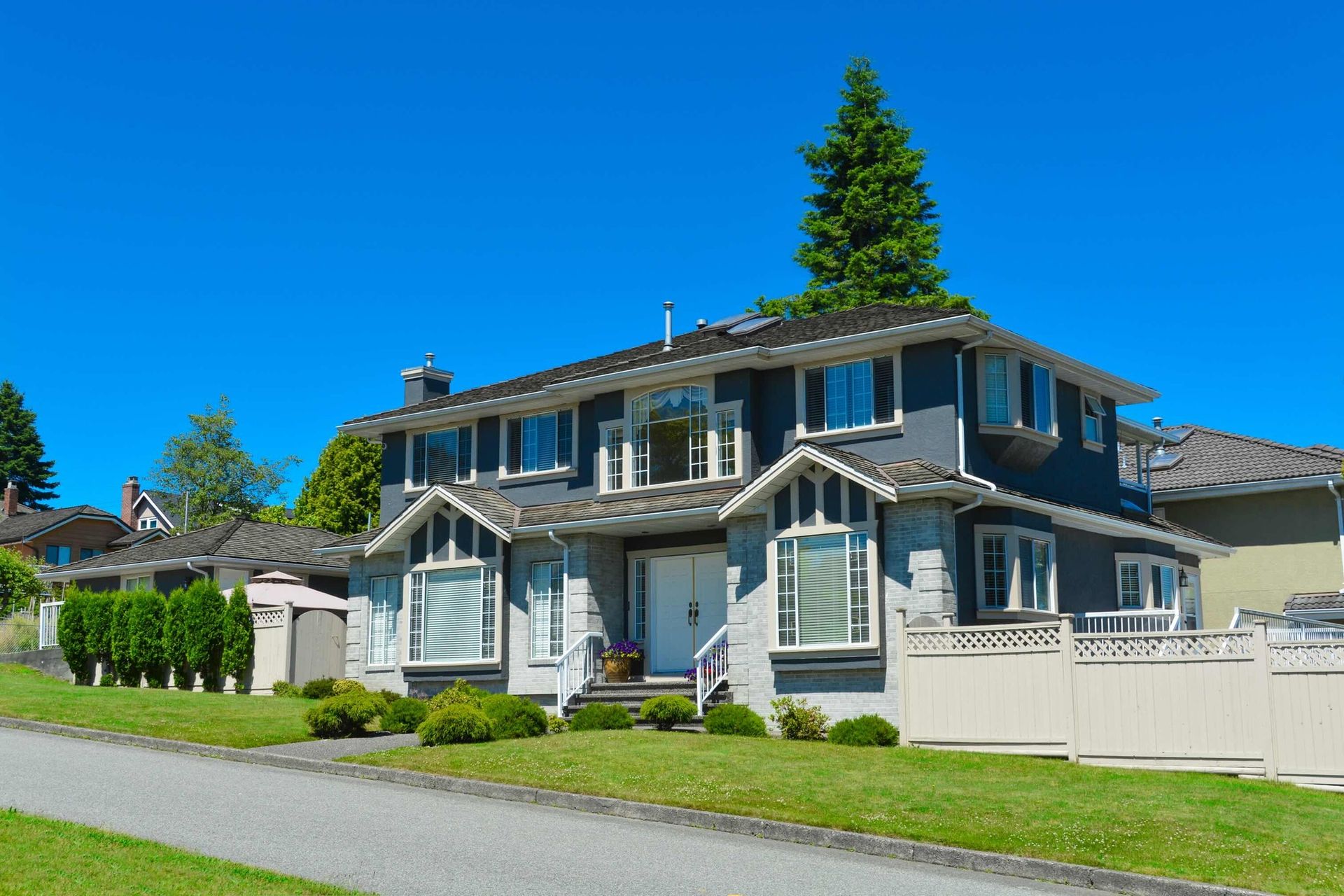 Two-story blue-gray suburban house with white trim and a tall evergreen tree under a clear blue sky