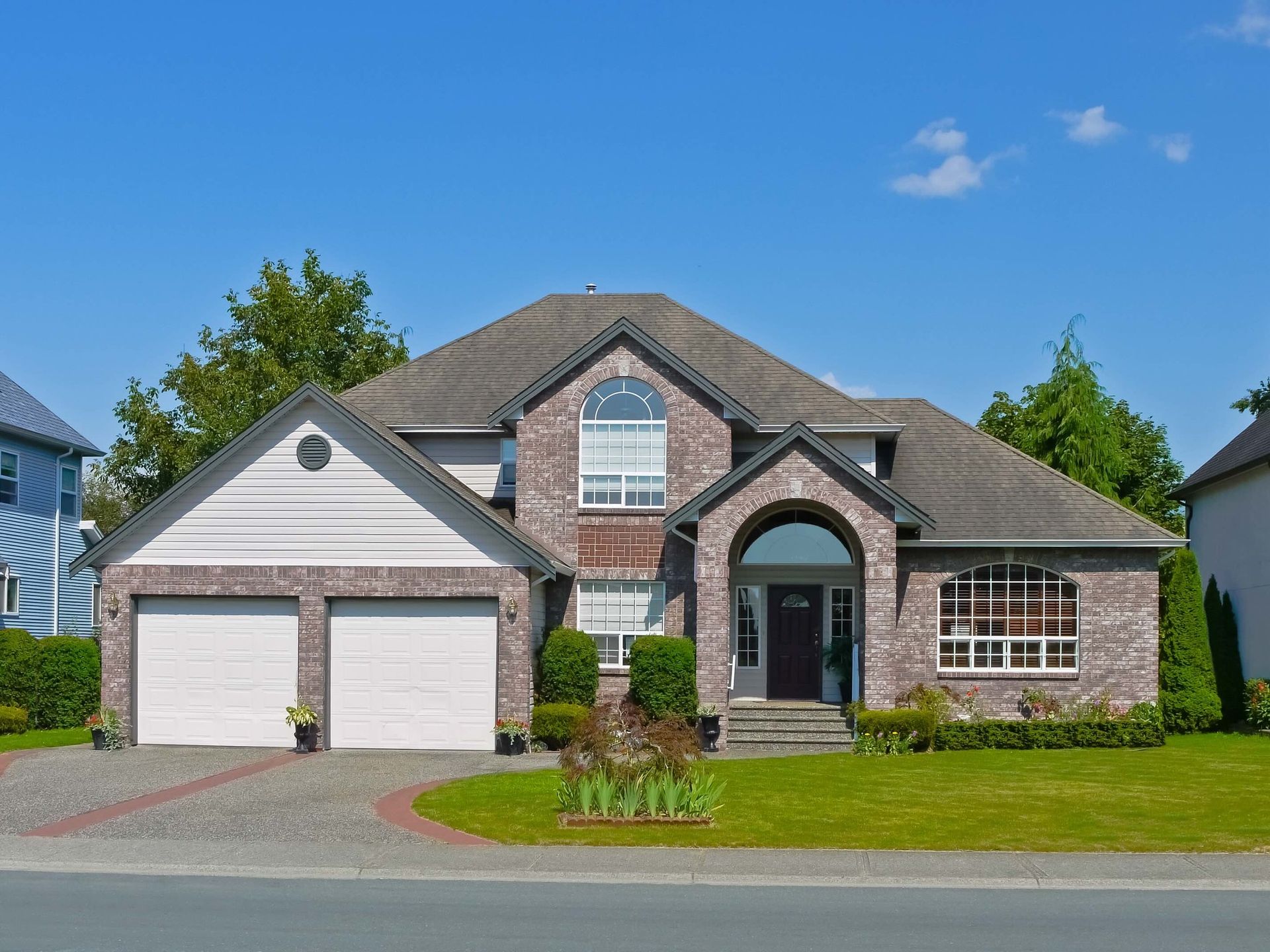 Suburban brick house with two-car garage, arched entry, and manicured lawn under a blue sky