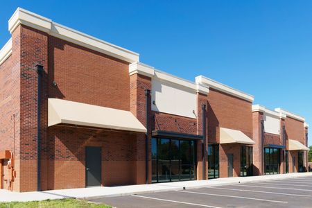 Empty brick storefronts in a strip mall with awnings and a parking lot under a blue sky
