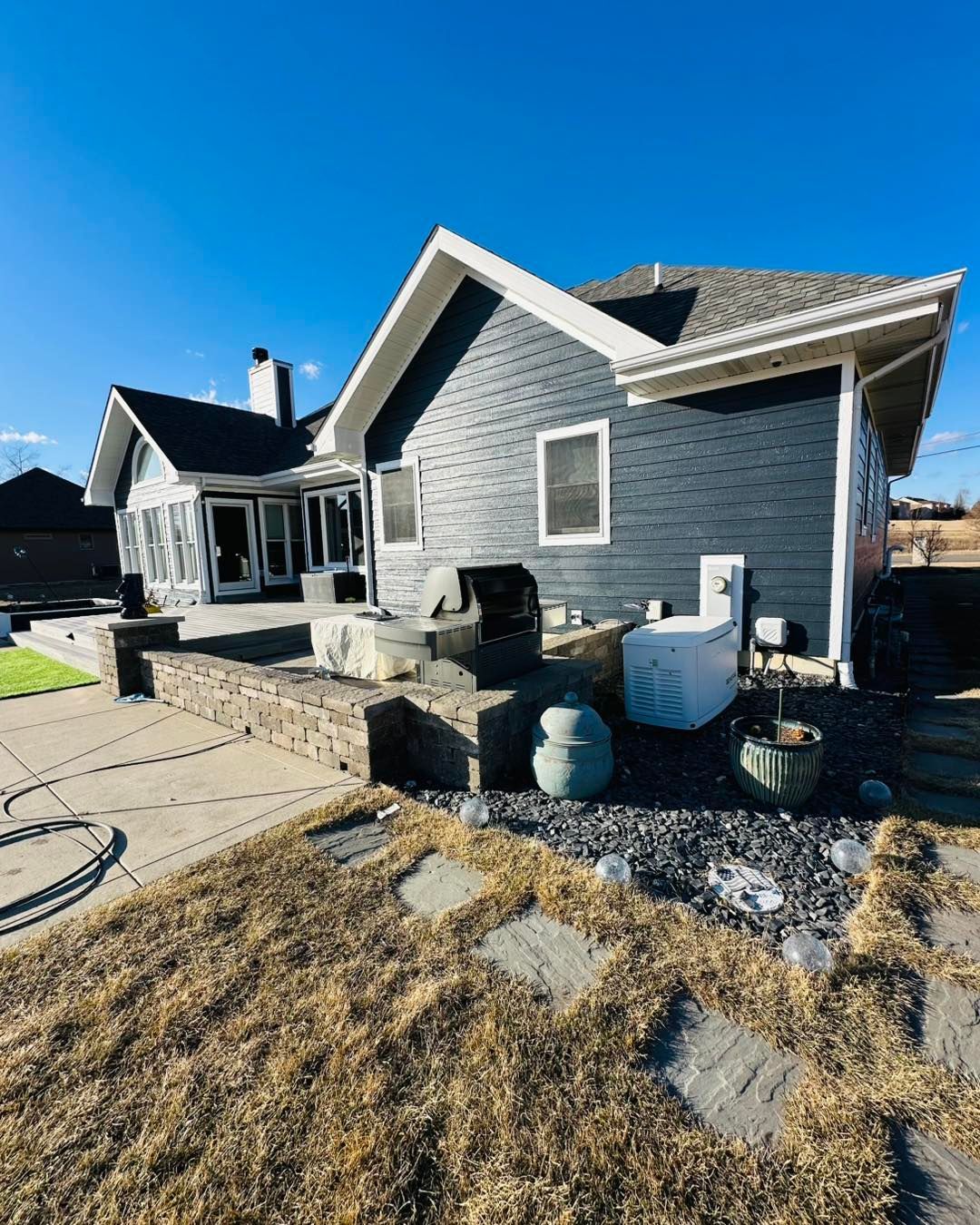 Gray house exterior with front patio, white trim, and blue water barrel under a clear blue sky