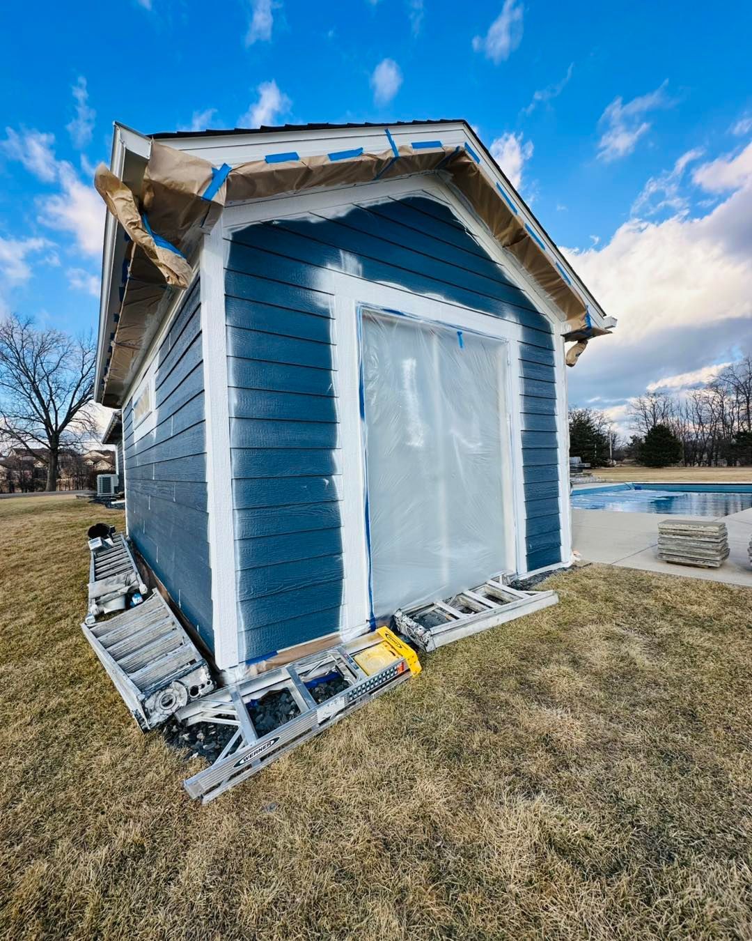 Blue house under construction with white trim, covered windows, and ladder on the side on a grassy lot