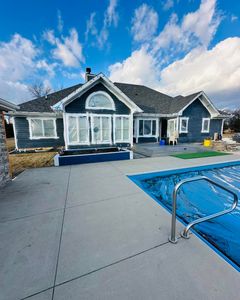 Blue-gray house with white trim beside a backyard pool and concrete patio under a partly cloudy sky