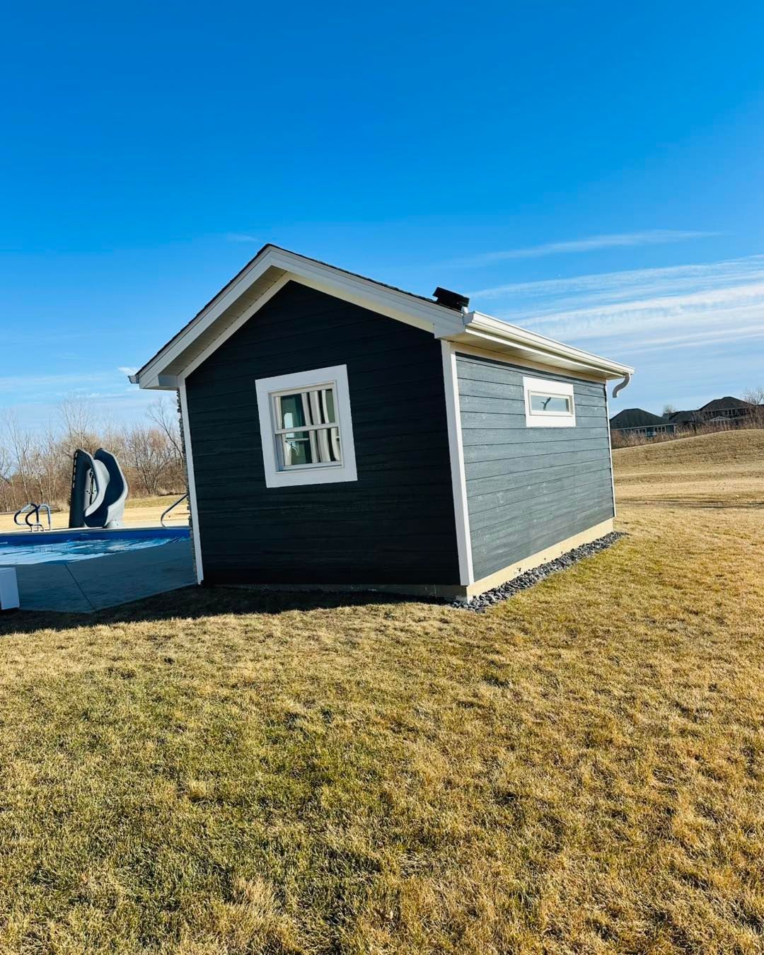 Small dark shed with white trim on a grassy field under a clear blue sky.