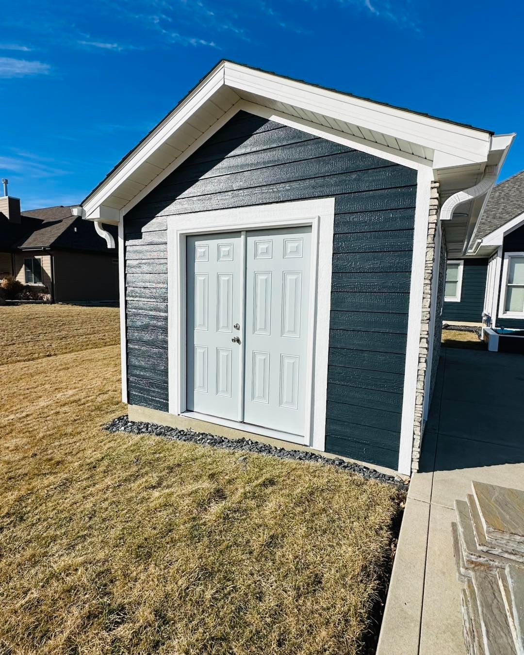 Small gray shed with white double doors and a gabled roof on a grassy lawn under a blue sky