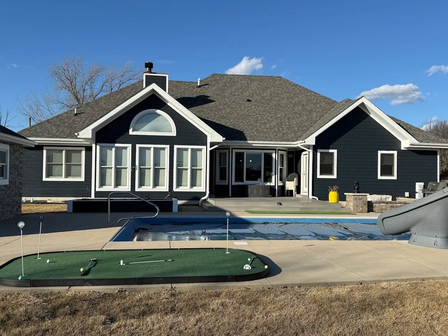 Single-story dark house with blue-trimmed windows and a large backyard pool under construction in front.