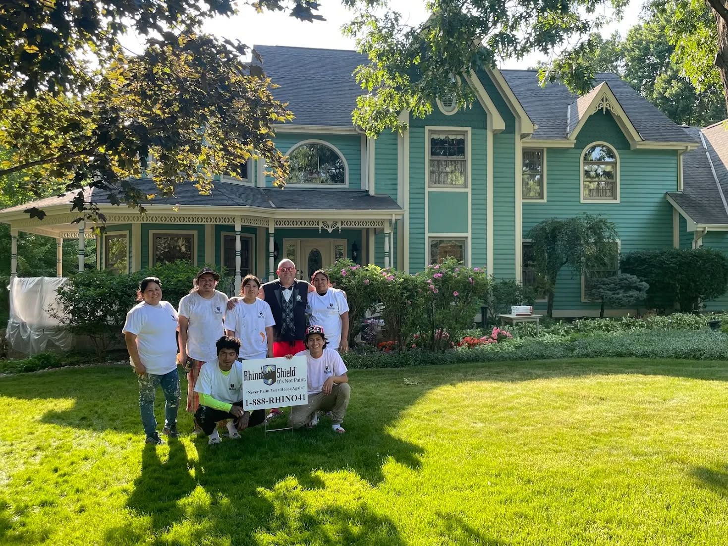 Group posing on a lawn in front of a large green house with a porch and flowers.