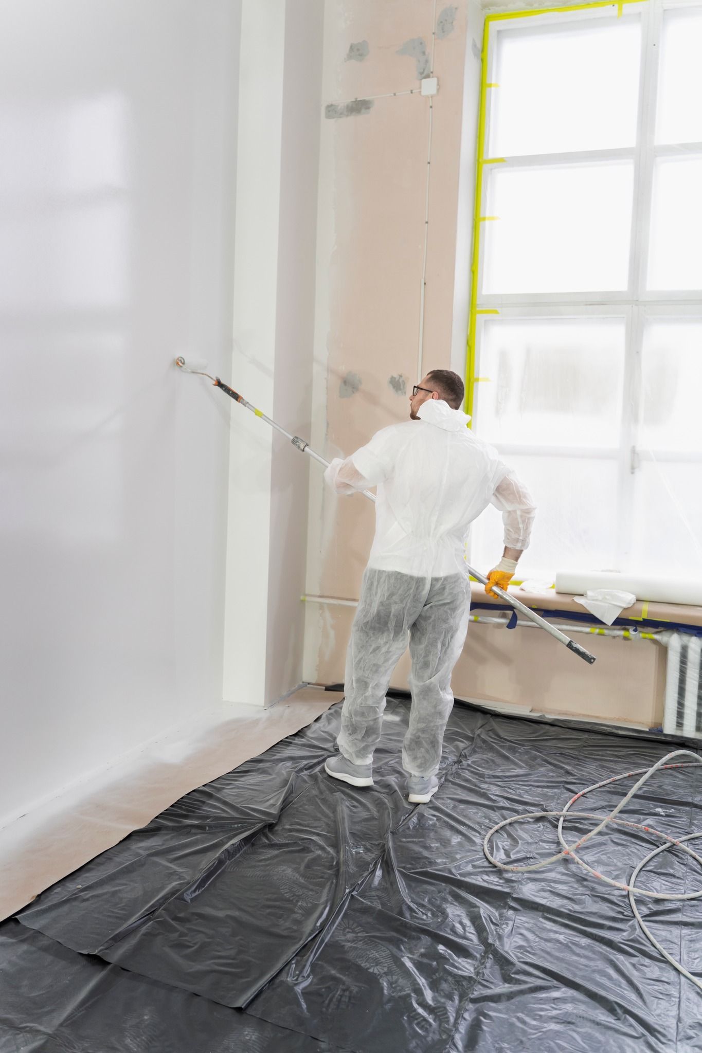 Person painting a wall in a bright room, standing on a plastic-covered floor near a window.