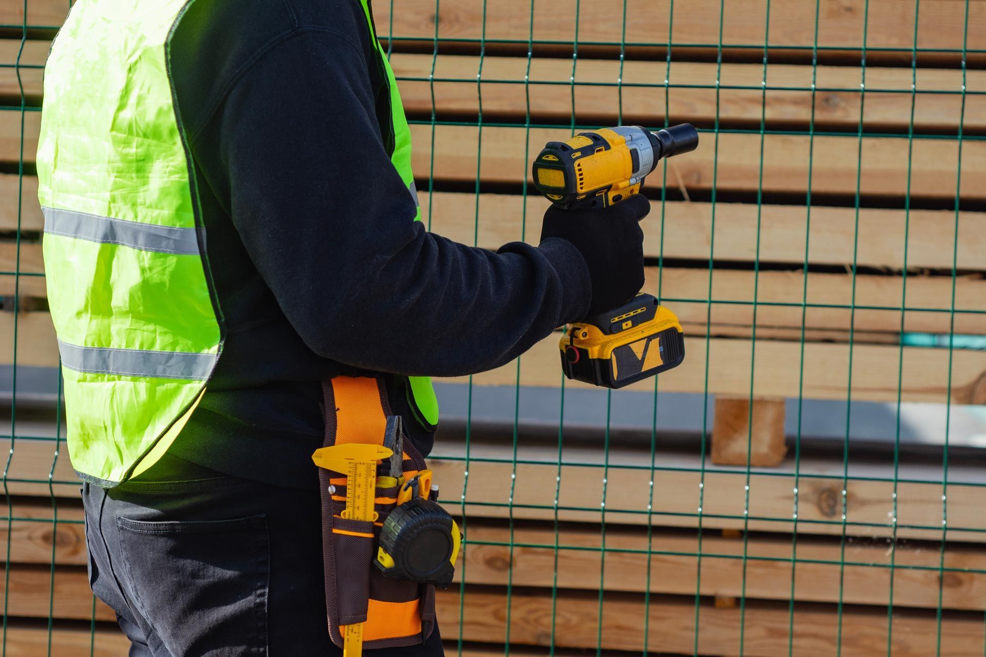 Worker in a safety vest holding a cordless power tool beside a wire fence and stacked lumber.