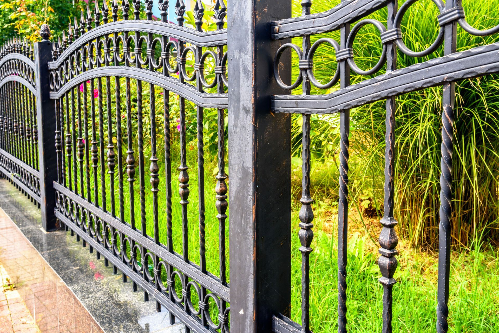 Decorative black iron fence with curved details lining a lush green garden walkway. Decorative black iron fence with curved details lining a lush green garden walkway.