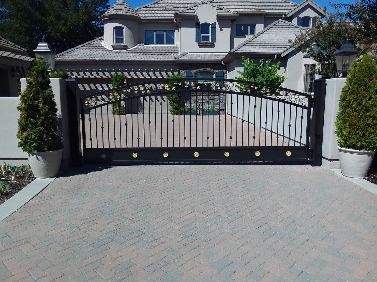 Black wrought-iron driveway gate before a large stucco house, flanked by potted topiary trees