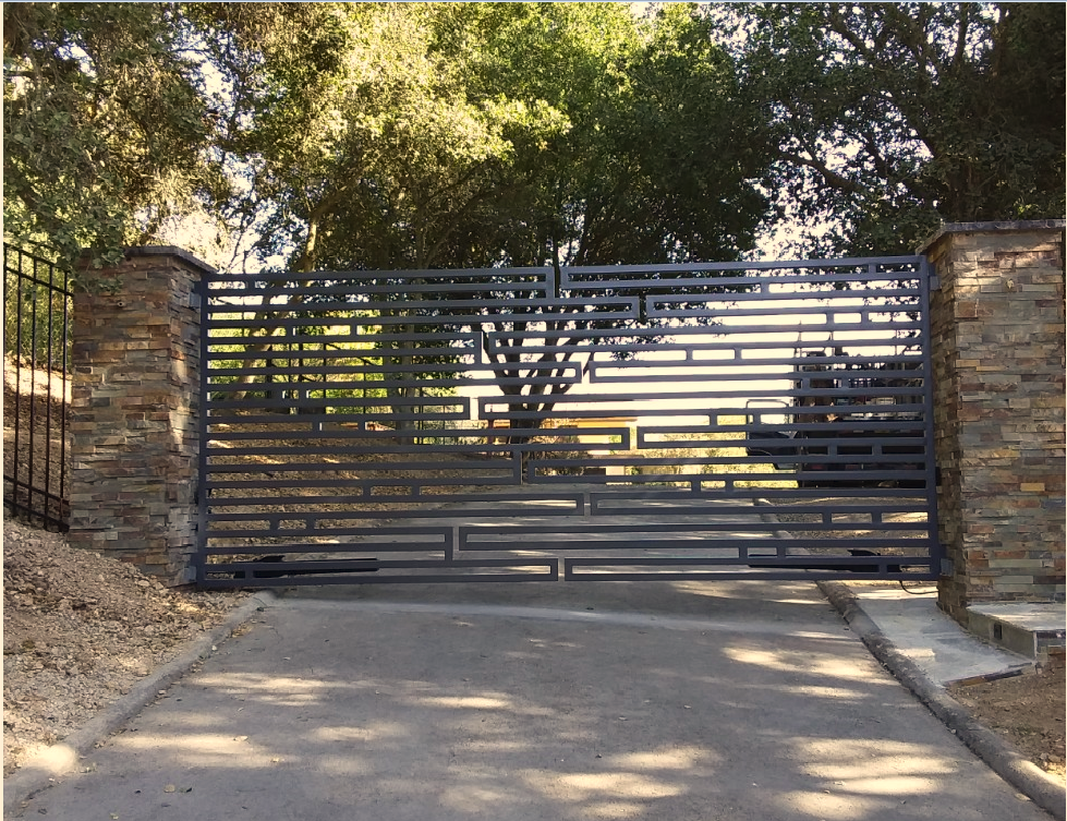 Black metal driveway gate between stone pillars, with trees and a paved road behind it