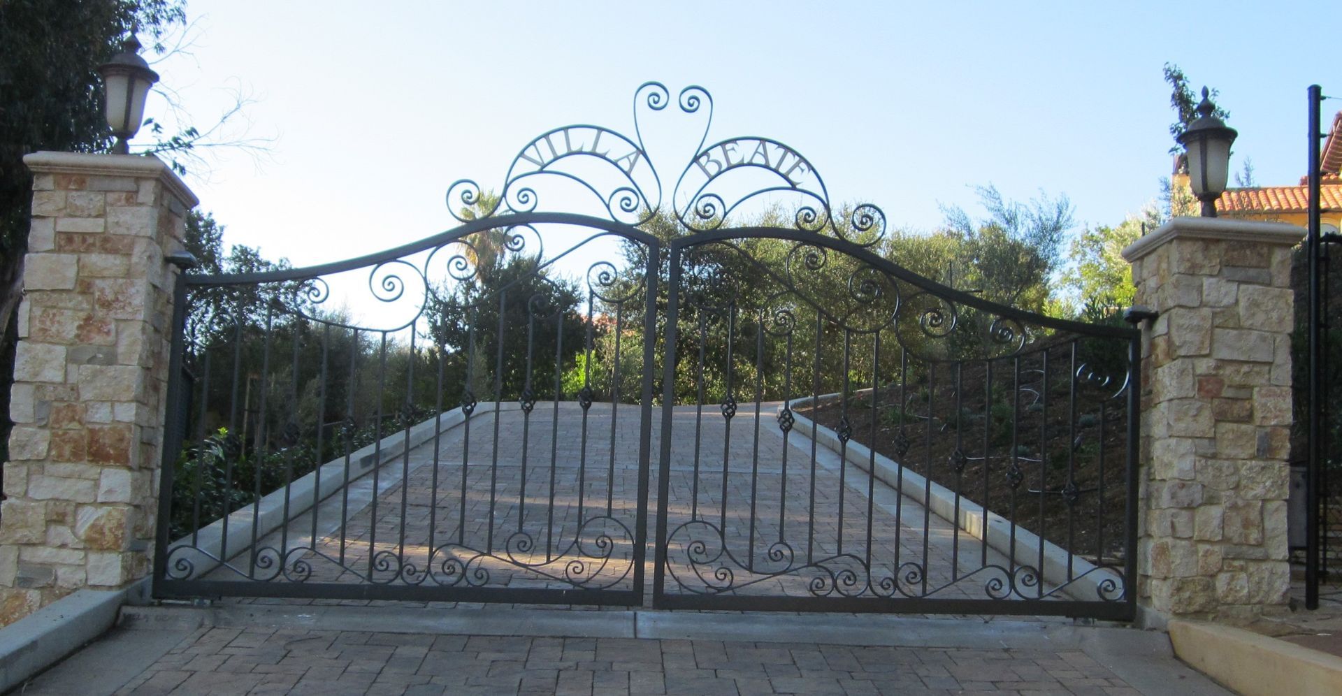 Ornate wrought iron gate with stone pillars; driveway in a natural setting.