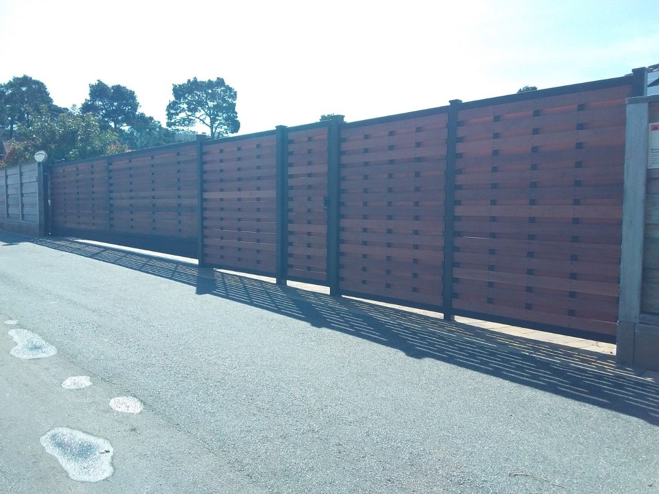 Long brown wooden fence lining a sunlit roadside with trees in the background