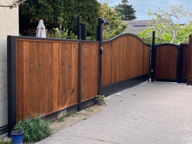Wooden sliding gate and fence along a paved driveway, with black metal posts and green trees nearby