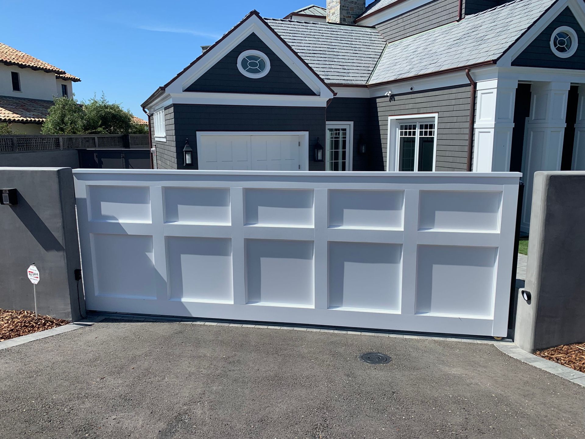 White paneled driveway gate in front of a modern home with gray siding. White paneled driveway gate in front of a modern home with gray siding.