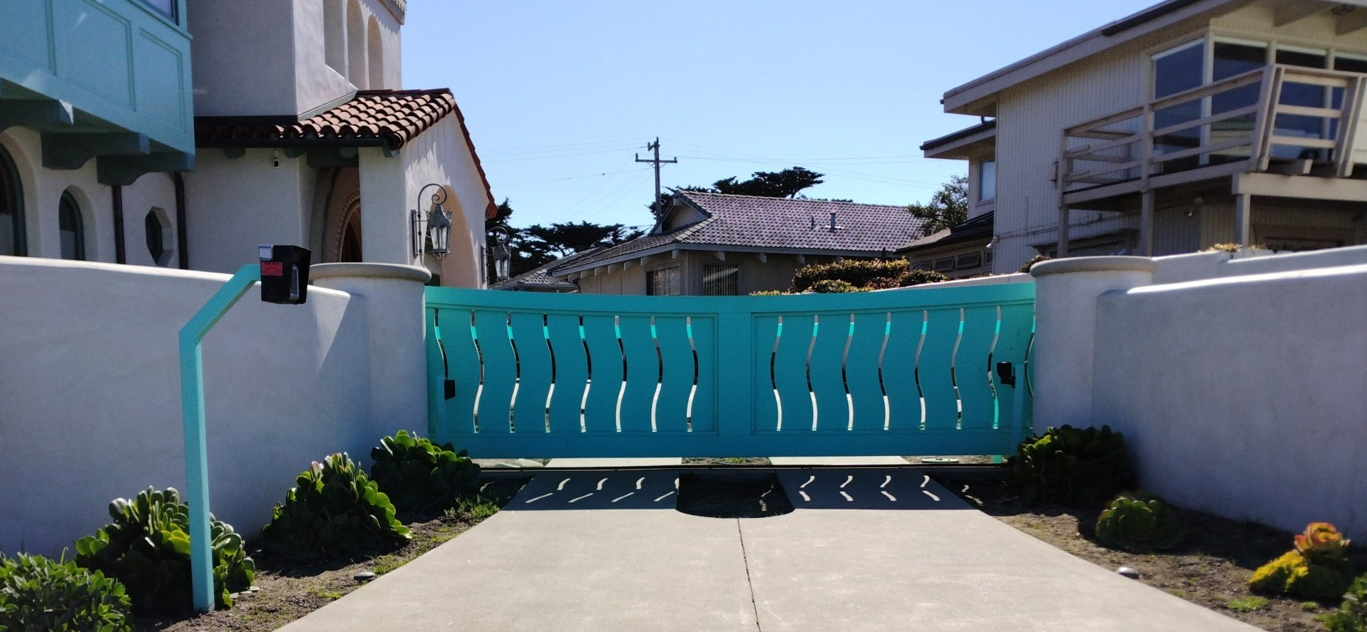 Turquoise gate across a driveway between white walls, with houses and greenery in the background.