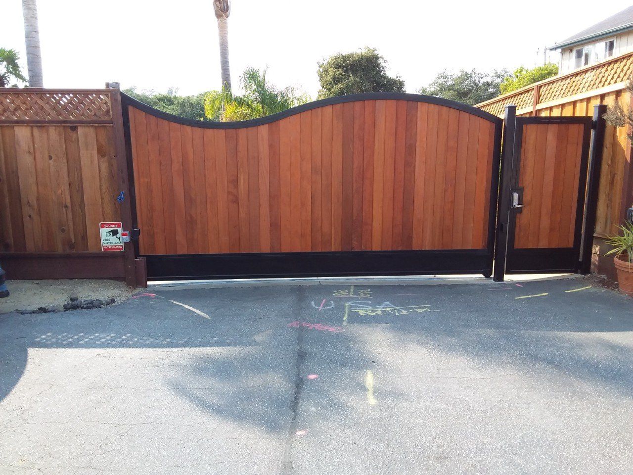 Wooden driveway gate with curved top panels and black metal posts beside a paved street