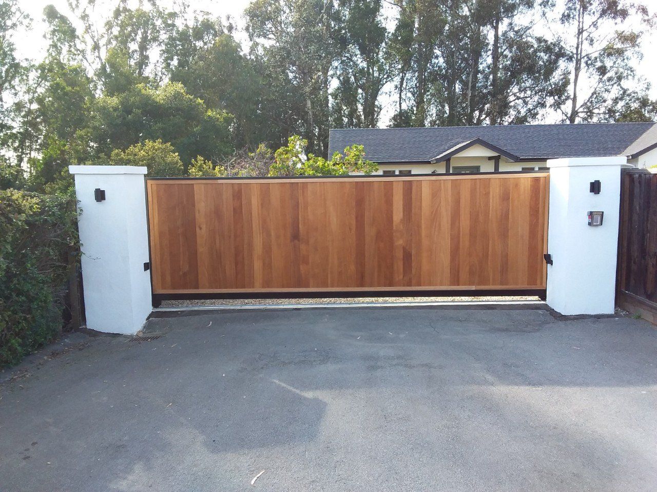 Wooden sliding gate between white pillars at a driveway entrance with trees behind it