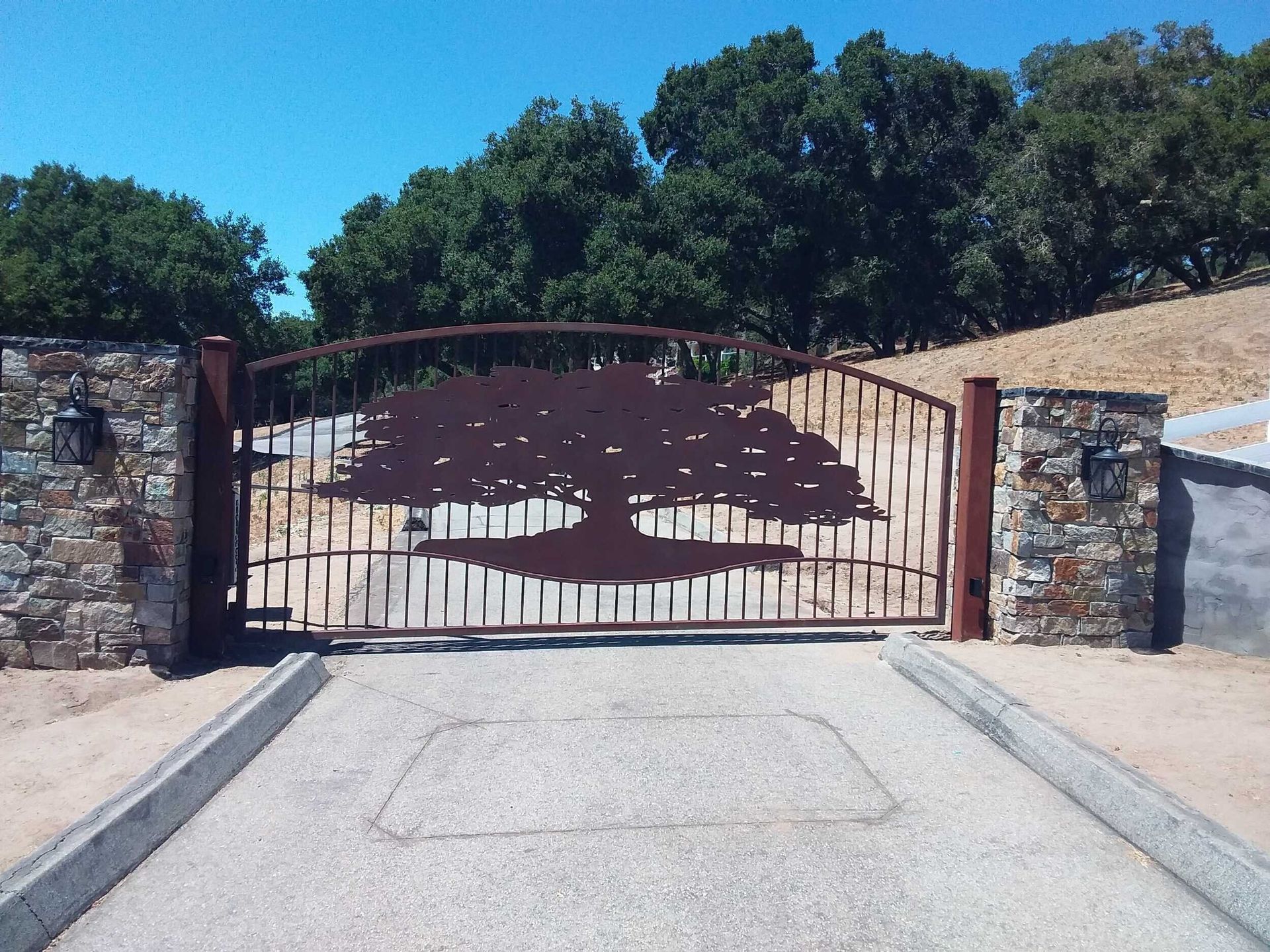Closed metal gate at a stone driveway entrance with trees and blue sky beyond