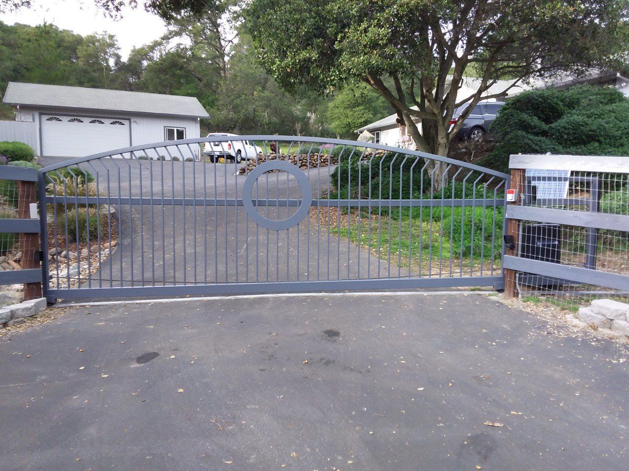 Closed metal driveway gate at a residential entrance, with a paved driveway and trees behind it