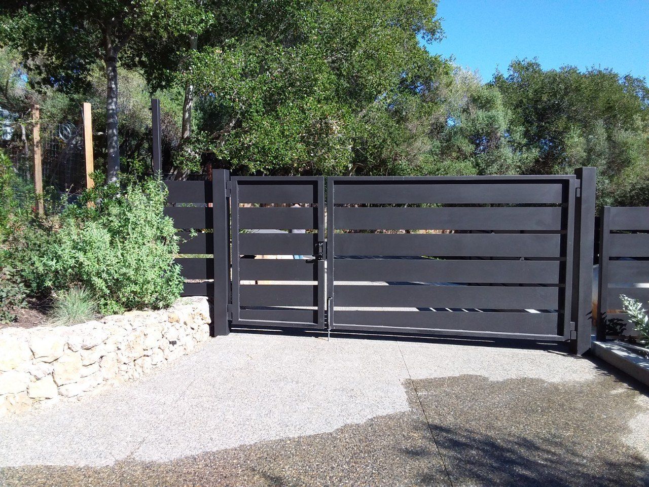 Black metal driveway gate beside a stone wall and gravel path, surrounded by greenery.