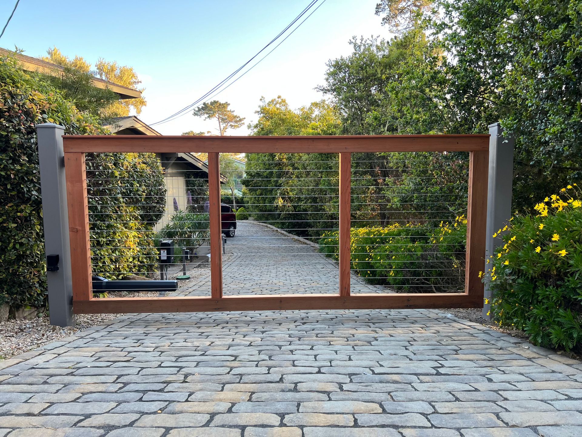 Wooden sliding gate across a paved driveway, framed by gray pillars and greenery beyond