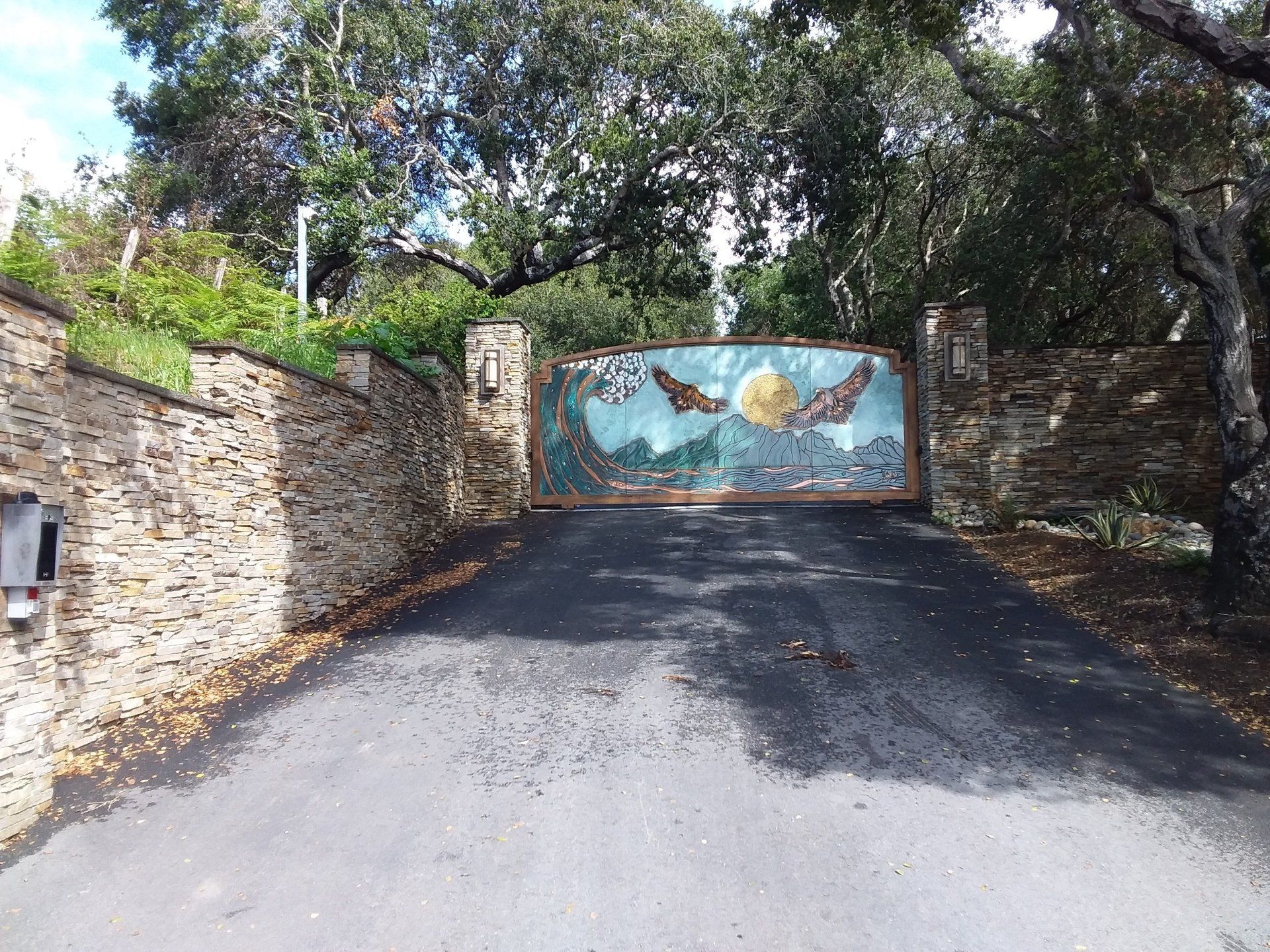 Driveway leading to a stone gate with a painted mural, framed by trees and stone walls.