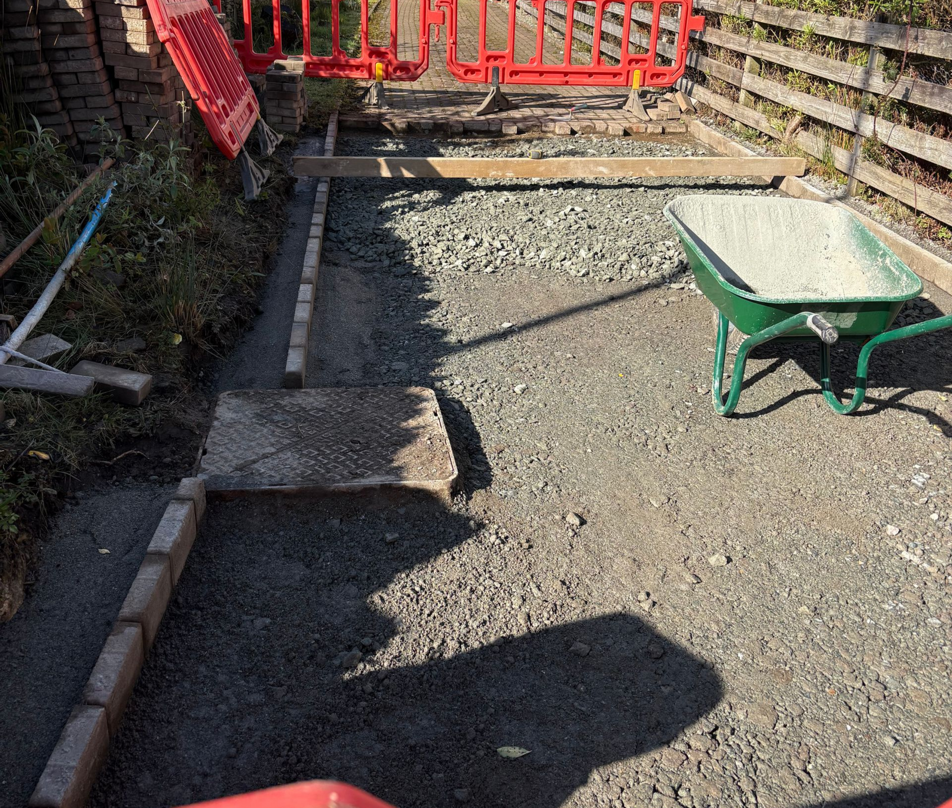 Gravel-covered backyard with red gate, stone borders, and a green wheelbarrow on the right